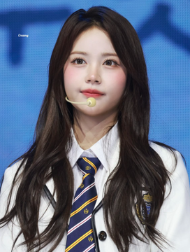A young Asian woman with long dark wavy hair, wearing a headset microphone, white shirt, and a blue and yellow striped tie, posing against a blue background.