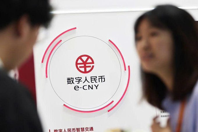 A blurred photograph of a display for the digital Chinese currency (e-CNY), featuring the red circular logo and Chinese text "数字人民币" and "e-CNY", with people standing in the foreground out of focus.