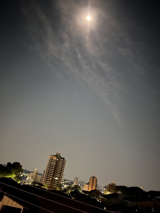 A nighttime cityscape photograph showing a bright moon in a hazy sky above illuminated buildings and a dark rooftop in the foreground.