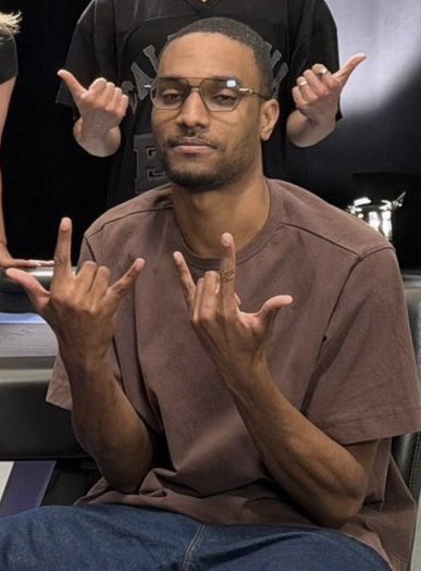 A man wearing glasses and a brown t-shirt sits with a neutral expression while making hand gestures in the foreground, with another person behind him mirroring similar poses. The image accompanies the news of ANSEM's suggestion to convince Coinbase to host a "Crypto Twitter Olympics," with the hand gestures possibly symbolizing sports or athletic competition referenced in the announcement.