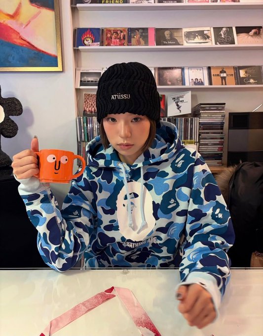 A person wearing a black "ATRISSU" beanie and a blue camouflage BAPE hoodie, holding an orange mug, seated in front of shelves filled with CDs.
