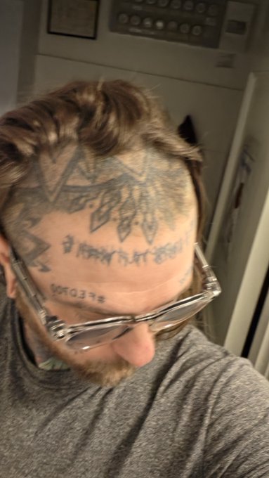 A person with tattoos on their forehead and scalp, wearing clear-framed glasses and a gray shirt, is photographed from a close, slightly blurry angle.