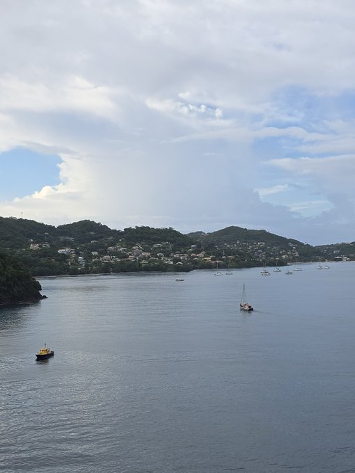 An aerial view of a coastal bay with boats anchored in calm water, with green hills and houses visible in the background under a partly cloudy sky.