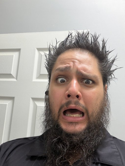 A close-up selfie of a man with a wild beard and spiked hair making an exaggerated shocked expression in front of a white paneled door.