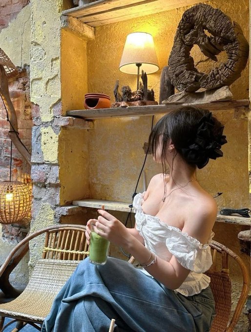 A woman with a black hair scrunchie sits in a rattan chair against a yellow textured wall, sipping a green beverage while a lamp and carved wooden sculpture sit on the shelf above her. The cozy, contemplative "coffee first" scene complements the post's message about taking a slow, thoughtful approach to trading rather than rushing into the noisy market.