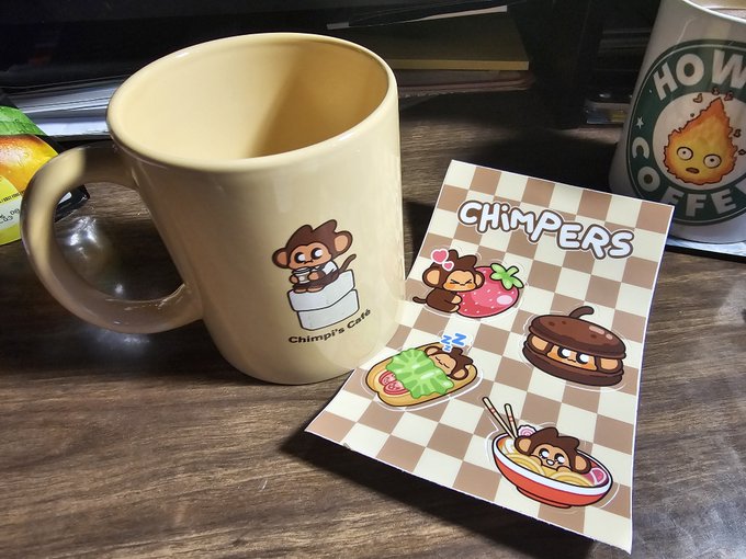A Chimp's Café branded mug sits next to a sticker sheet featuring cute cartoon monkeys with various food items like strawberries and ramen.