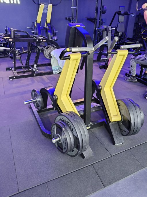 A yellow plate-loaded fitness machine loaded with weight plates sits in a gym with blue lighting.