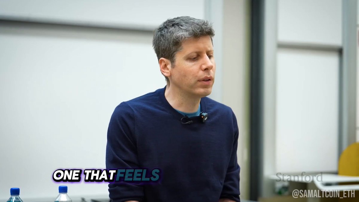 The image features a man wearing a blue sweater sitting at a desk with two water bottles next to him. The background of the image shows a white wall, which serves as a backdrop for the text "One That Feels" written on it. This phrase is likely part of a caption or description related to the content displayed in the image.