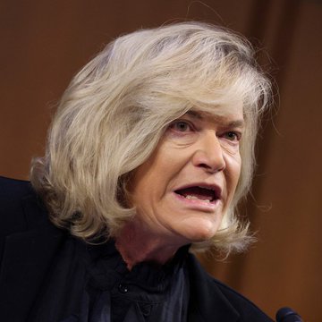 urn of water, a woman with blonde hair and blue eyes, dressed in black, and a brown wall backdrop.