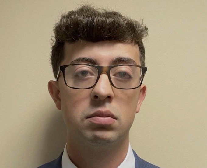 A close-up portrait shows a young man with curly brown hair, thick dark-rimmed glasses, and a neutral expression, wearing a white shirt against a plain beige background.