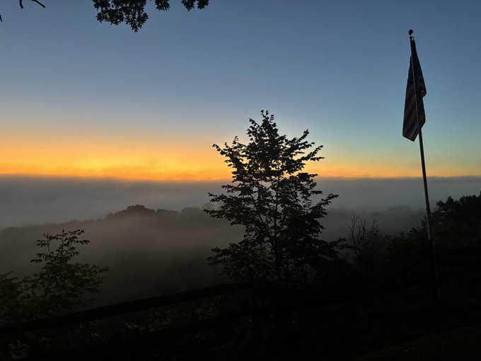 A serene sunrise over a fog-filled valley features a silhouetted tree and an American flag on a flagpole in the foreground.