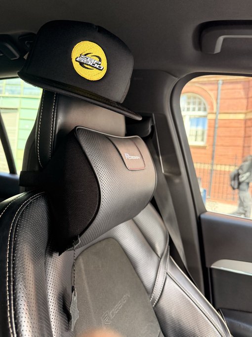 An interior photo of a car showing the front seat with a black neck pillow and a yellow Lakers cap hanging on the headrest.