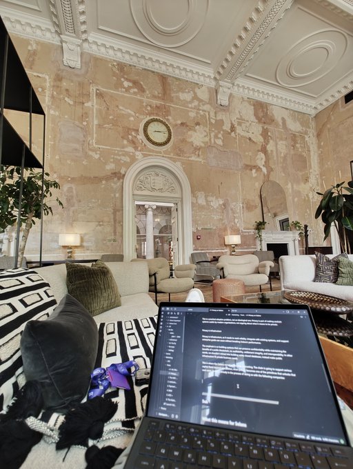 A laptop displaying text/code sits on a couch in an elegant room featuring ornate ceiling moldings, weathered plaster walls, a round clock, and classic furnishings.