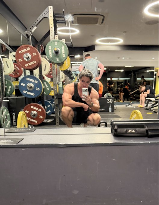 A muscular man in a black tank top takes a mirror selfie in a gym, crouching down with his phone while squat racks loaded with Eleiko weight plates are visible in the background.