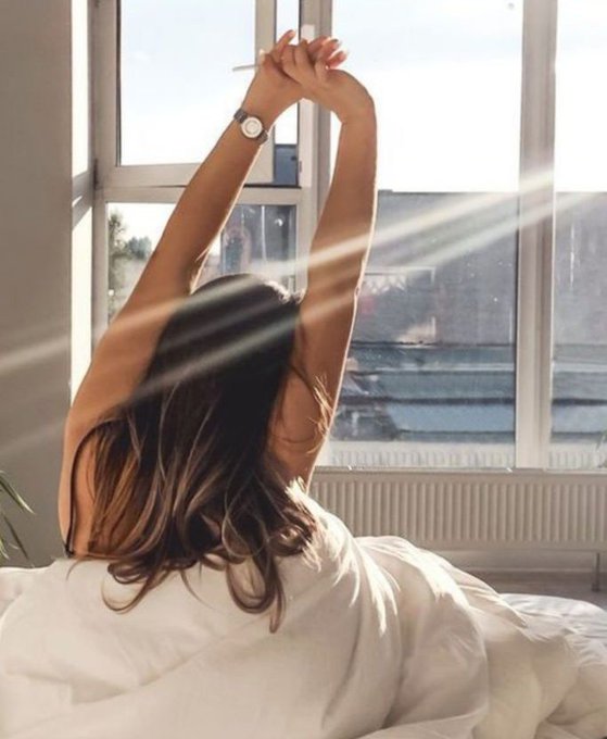 A woman with long brown hair stretches her arms above her head while sitting on a bed, sunlight streaming through a large window behind her.