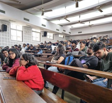 A diverse group of young students sits attentively in a tiered university lecture hall, with a woman in a red sweater prominently featured in the foreground and others engaged with devices or conversation. The scene captures the engaged atmosphere of the #POCIndiaOnChainTour, reflecting the post's message of welcoming "curious minds" to learn about blockchain technology across Indian campuses.