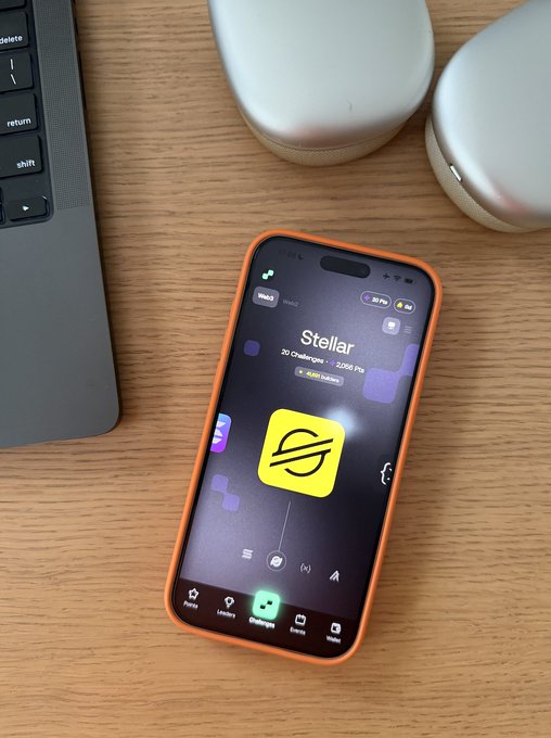 The image features an orange smartphone placed on top of a wooden table, with its screen displaying the app "@StellarOrg" along with some other icons. The background shows a laptop computer next to the phone, suggesting that someone might be using both devices for work or leisure purposes.