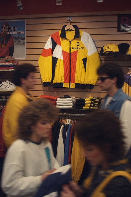 A retro-styled yellow, red, and white color-blocked track jacket bearing the "BINANCE" logo hangs prominently in a vintage sportswear shop, surrounded by shoppers and racks of athletic apparel. The bold, graphic design evokes classic 80s-90s sports fashion, supporting the claim that its timeless, ahead-of-its-time fit still feels fresh today.