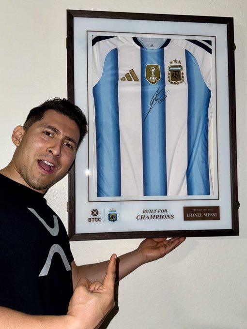 A man poses excitedly next to a framed signed Argentina national team jersey with "BUILT FOR CHAMPIONS" and "LIONEL MESSI" displayed at the bottom, pointing toward the plaque. The image is unrelated to the XRP giveaway post, as it simply shows a person celebrating a sports memorabilia piece with no cryptocurrency or text related to the promotion visible.