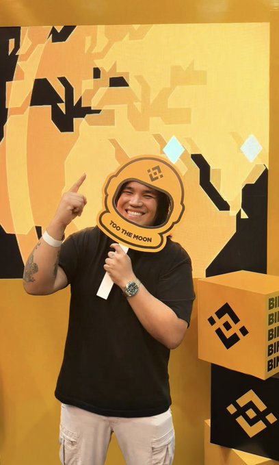 A man wearing a black t-shirt smiling and pointing at a cardboard cutout of the Binance logo on a stick that reads "TO THE MOON," standing next to a large yellow Binance cube logo.