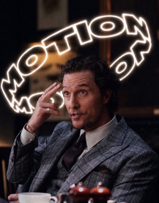A man in a gray plaid suit sitting at a table with a neon sign reading 'MOTION' reflected behind him.