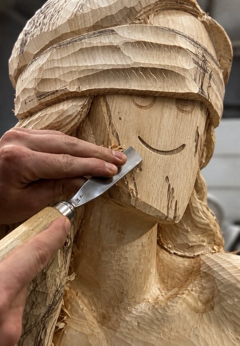 A close-up shows hands using a chisel to carve fine details into a wooden sculpture of a smiling figure wearing a head covering.