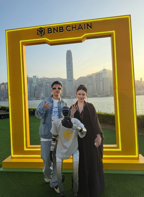 Two people pose with a humanoid robot in front of a large yellow "BNB CHAIN" frame, with Hong Kong's skyline visible in the background, celebrating the BNB Chain's 6th anniversary milestone. The image ties to the post's reflection on attending BNB Chain events, highlighting community engagement and the ecosystem's growth over the past six years.
