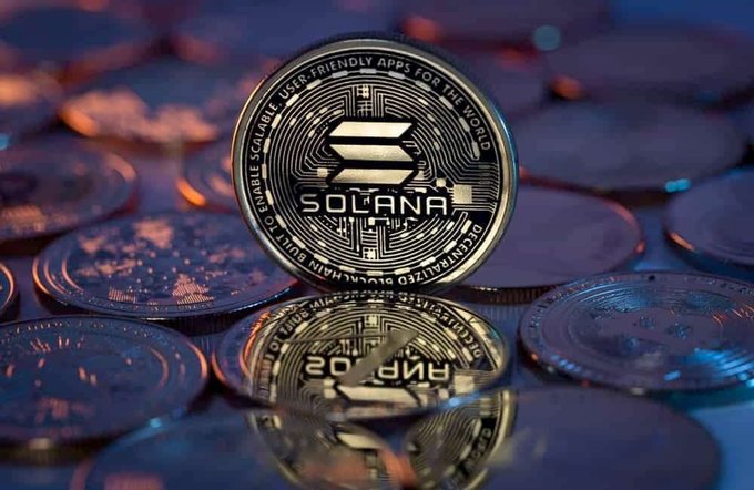 A close-up photo of a silver Solana cryptocurrency coin standing upright on a reflective surface with other coins in the background.