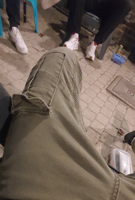 A close-up shot shows a person wearing olive green cargo pants standing on a paved outdoor surface near others wearing white sneakers.