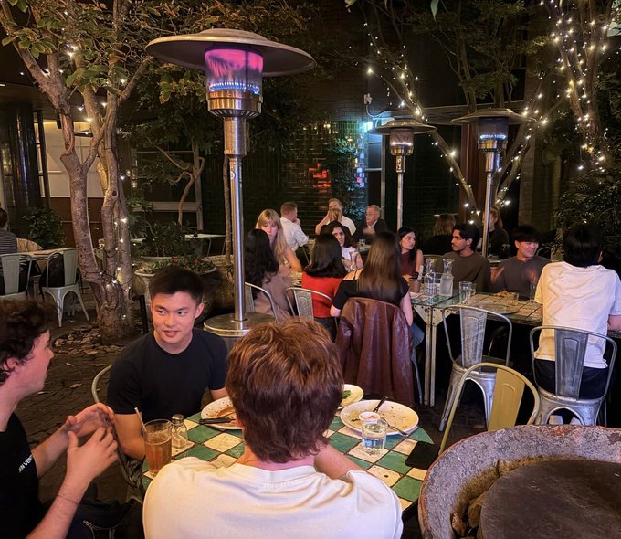 A group of people dining at an outdoor patio restaurant at night, with patio heaters illuminating the scene.