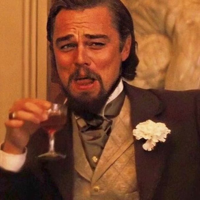 Leonardo DiCaprio, dressed in period clothing with a white carnation, holds a glass of red wine with a confused or disgusted expression.