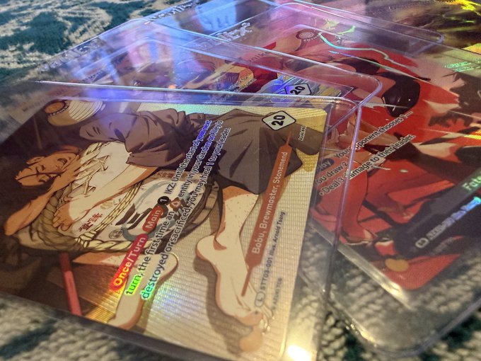 A close-up of holographic Azuki TCG cards in protective sleeves, with the visible "20" Bobu character card showing the textured and foiled leg hair details highlighted in the post. The other cards in the background display additional artwork and text, including phrases like "turned," "I turn," and "Once up."