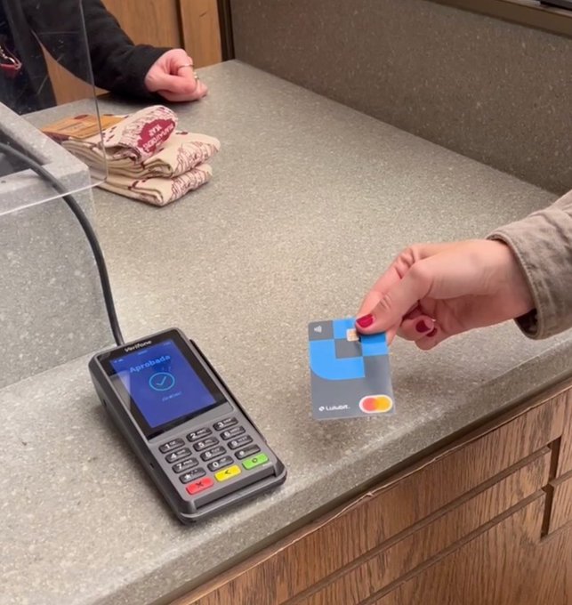 A person holds a Walmart Mastercard credit card near a Verifone payment terminal on a counter, with another person's hands visible in the background.