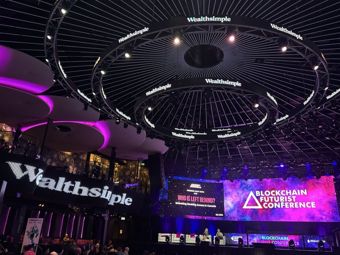 The image captures the Main Stage of the Blockchain Futurist Conference, prominently featuring Wealthsimple branding displayed across large circular ceiling installations and on screen, reflecting their major sponsorship role at the event. A panel discussion appears to be underway, with the digital backdrop showing the event title and a session titled "WHO IS LEFT BEHIND?" addressing Redefining Wealth Access in Canada.