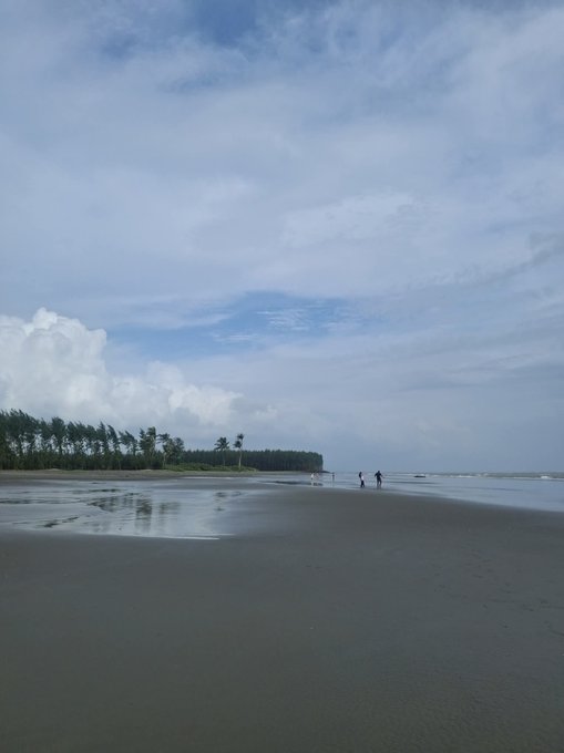 A serene beach scene at low tide under a partly cloudy blue sky, with two small figures walking along the wet sand near a line of wind-swept trees in the distance.