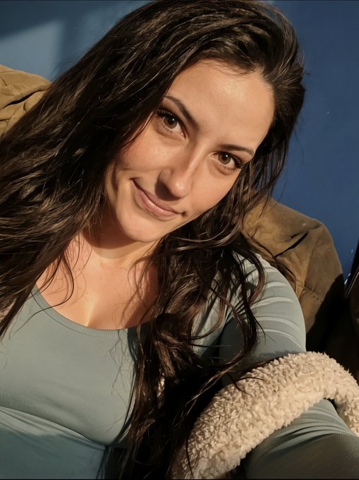 A selfie-style photo of a young woman with long dark wavy hair wearing a light blue/grey top, smiling softly at the camera with warm lighting.