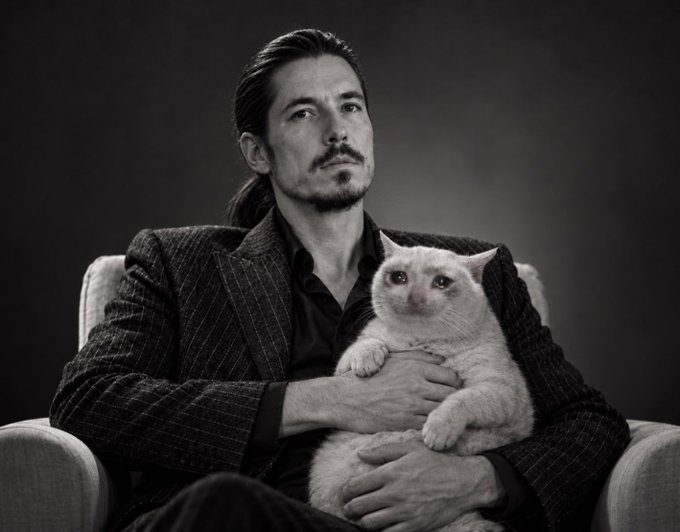 A black and white portrait shows a man with long dark hair and a mustache, wearing a pinstripe suit, holding a light-colored cat in his arms while seated.