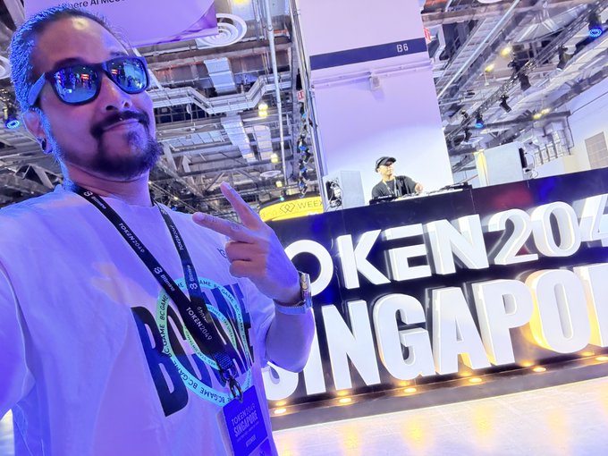 A man wearing sunglasses and a "Token2049 Singapore" lanyard strikes a peace sign in front of a large illuminated "TOKEN 2049 SINGAPORE" sign, embodying a "GM fam" greeting to fellow crypto enthusiasts at the prominent Web3 conference.