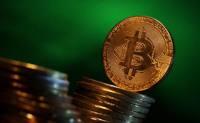 A physical Bitcoin token rests atop a stack of gold coins against a green backdrop, visually representing the surge of capital into spot Bitcoin ETFs—particularly BlackRock's fund—following Monday's $999 million inflow, the largest daily intake since October 2025. The engraved text "BITCOIN DIGITAL DECENTRALIZED PEER TO PEER" around the coin's edge reinforces the cryptocurrency's role as the asset driving this renewed institutional demand.