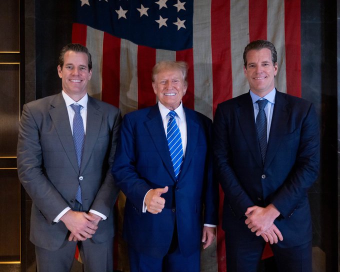 Donald Trump giving a thumbs-up while standing between his two sons, Donald Trump Jr. and Eric Trump, in front of an American flag.