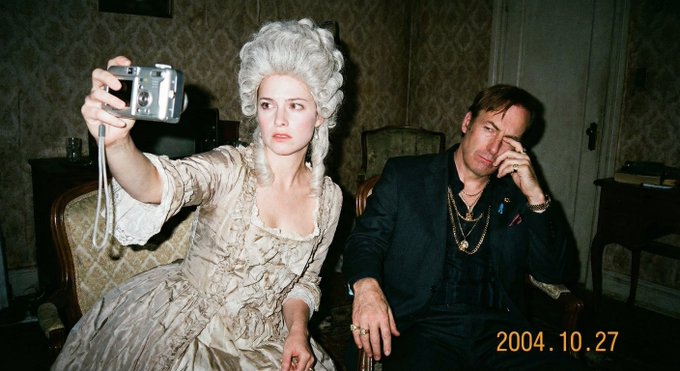 The image features a man sitting next to a woman who is taking his picture with her camera. The man appears to be wearing a suit, while the woman has blonde hair and is dressed in a white dress. They are both seated on chairs within an indoor setting that includes a couch and a dining table nearby.