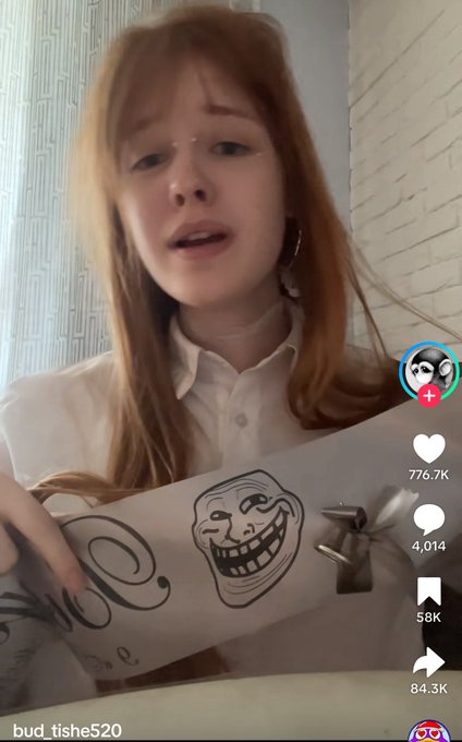 The image shows a woman holding up her arm, displaying a paper skateboard with an artistic and humorous design featuring a cartoon character's face. The word "trollface" can also be seen written above this figure. In the background, there is a text box that reads: "trollface still cooking after 18 years." This may seem unrelated to the image of the woman with the skateboard at first glance, but it might have been a reference or an inside joke between friends or someone close to her about the original post featuring the "trollface" character.