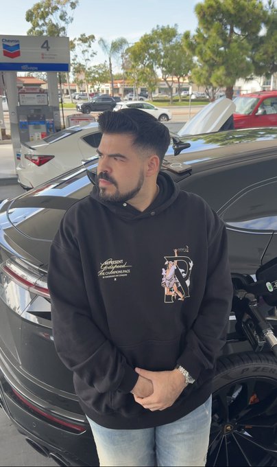 A man with a beard and styled hair wearing a black graphic hoodie, standing next to a black sports car at a Chevron gas station.