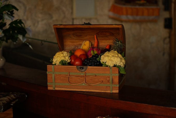 An open wooden treasure chest filled with fresh produce including cauliflower, tomatoes, corn, carrots, grapes, and an orange sits on a polished dark wooden table.