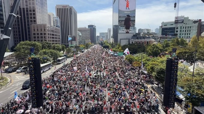 An aerial view shows a massive crowd of people filling a city street during what appears to be a large protest or rally, surrounded by tall buildings.