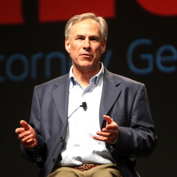 A man with gray hair, wearing a dark blazer and light blue shirt, speaks into a lapel microphone while gesturing with both hands on a stage with partial text reading "...corn... Ge..." visible in the background. While the image itself does not identify the speaker or show Greg Abbott, the accompanying post about Texas Governor Greg Abbott ordering a halt on data center permits suggests this may be related to his public discussion of energy grid and data center policy.