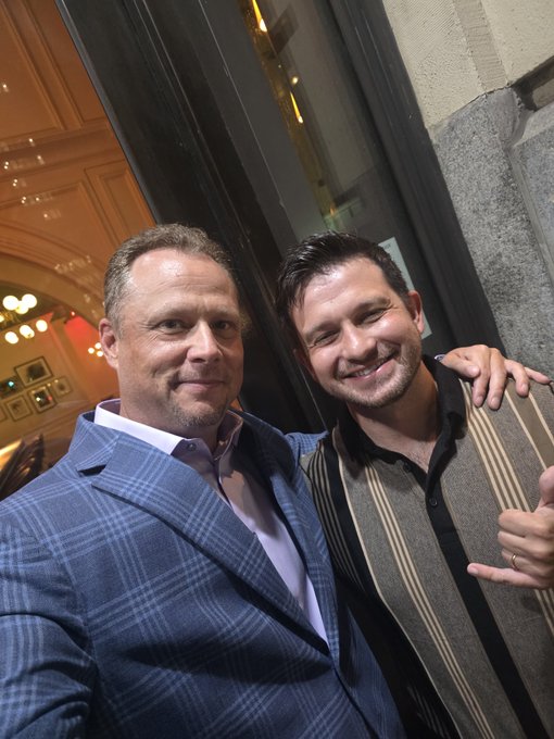 Two men pose closely together for a selfie inside what appears to be an ornate building; the man on the left wears a blue plaid suit, while the man on the right wears a striped shirt and vest.