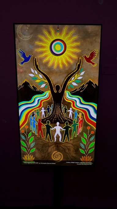 A vibrant painting featuring a silhouette figure with raised arms reaching toward a colorful sun, with people holding hands in a circle and rainbow ribbons flowing from the figure's base.
