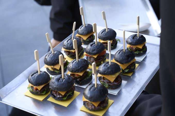 A server carries a tray of mini sliders featuring charcoal-black buns topped with melted cheese and lettuce, each secured with a wooden pick, presented under the "Burger N Buns" brand.