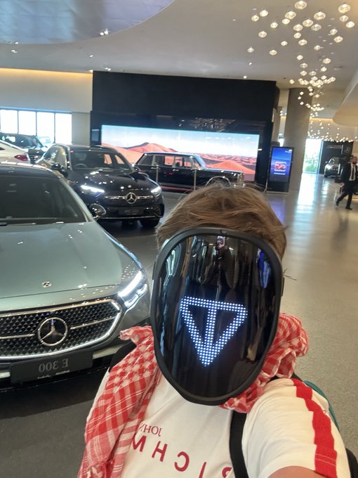 The image features a person wearing a face mask with a large "T" on their forehead, standing next to several cars at a car dealership. The person appears to be posing for a photo or selfie while surrounded by various vehicles. The caption of the image reads: "GM to the TON believers".